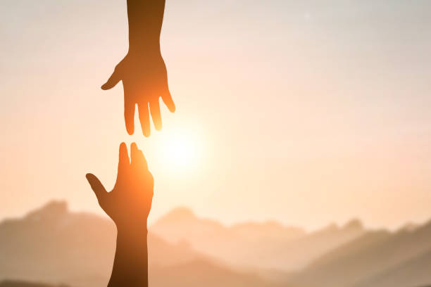 silueta de dos manos de personas que se alcanzan entre sí para ayudarse en el cielo del atardecer y el sol naranja. concepto de amistad, trabajo en equipo, ayuda, fe y esperanza. - ayuda fotografías e imágenes de stock