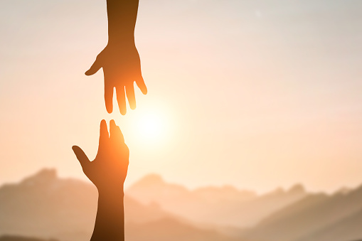 Silhouette of two people hands reaching to each other for help in sunset sky and orange sun. Friendship, teamwork, help, faith and hope concept.