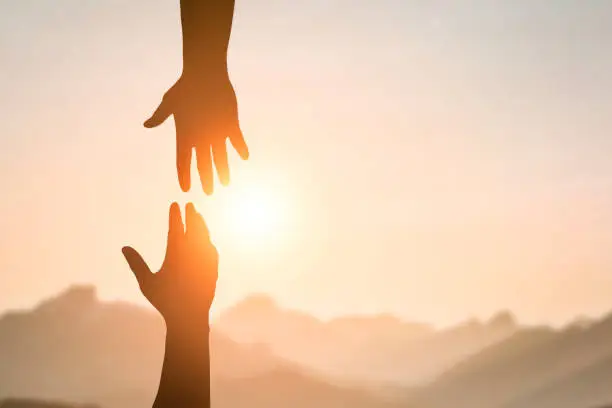Silhouette of two people hands reaching to each other for help in sunset sky and orange sun. Friendship, teamwork, help, faith and hope concept.