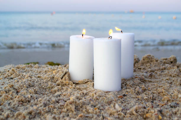 три большие белые парафиновые свечи горят на песчаном пляже на фоне моря - paraffin стоковые фото и изображения