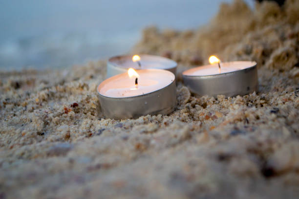 Three small burning candles on sand beach on background of blurry sea waves close-up. Candlelight, candle flame. Romantic mood backdrop. Concept date relaxation leisure vacation. Sandy shore sea coast