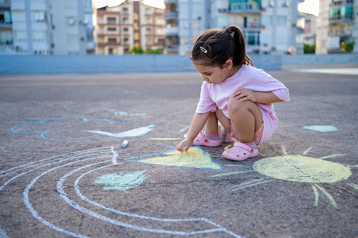 Cute girl drawing American flag with colored chalks on the sidewalk near the house on sunny summer day. Kids painting outside. Creative development of children. Patriotic day concept