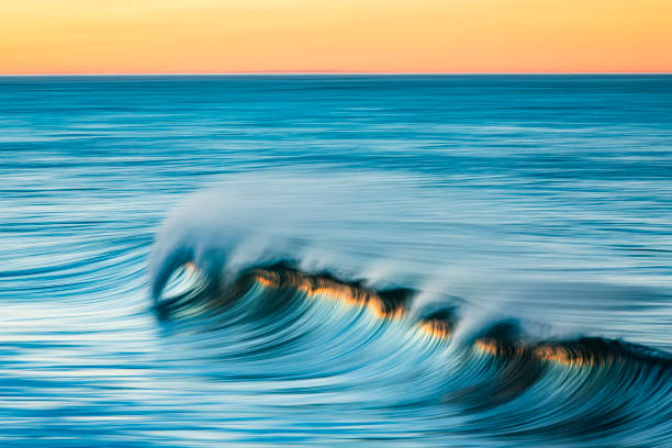 Smooth blue ocean wave breaking and reflecting a bright golden sunrise at the beach, Southern California, USA.