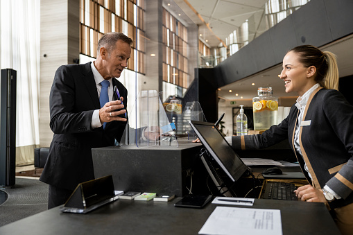 Mature businessman seen in a hotel during his business trip holding a smartphone with a booking app on the screen and showing it to the female receptionist.