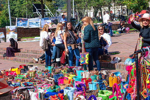 Bogotá, Colombia - May 21, 2017: The weekly Sunday event of the Mercado de Las Pulgas or Flea Market in the historic Usaquén District of the capital city, is in full swing. Some tourists look at brightly colored Mochila bags for sale. The focus of the Flea Market is on enjoyment: there is music, entertainment, shopping and a variety of food including various snacks and deserts, available. The altitude at street level is about 8,660 feet above mean sea level. Photo shot in the afternoon sunlight; horizontal format.