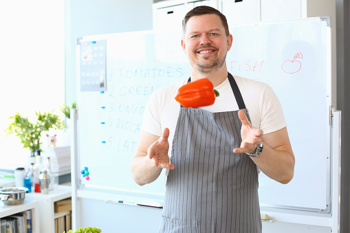 Portrait of smiling male cook catching flying red bell pepper. Man juggling vegetable. Cooking, preparing dish and making content for video blog concept