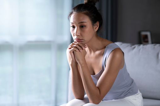 Asian woman feeling sad tired and worried suffering depression in mental health, Unhappy female sitting on couch at home, thinking about problems.
