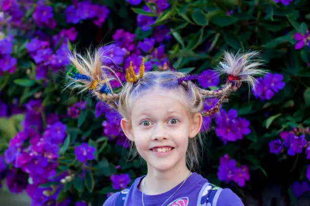 Photo of Funny little cute caucasian girl with braids for crazy hair day at school on purple flowers tibouchina nature outside background