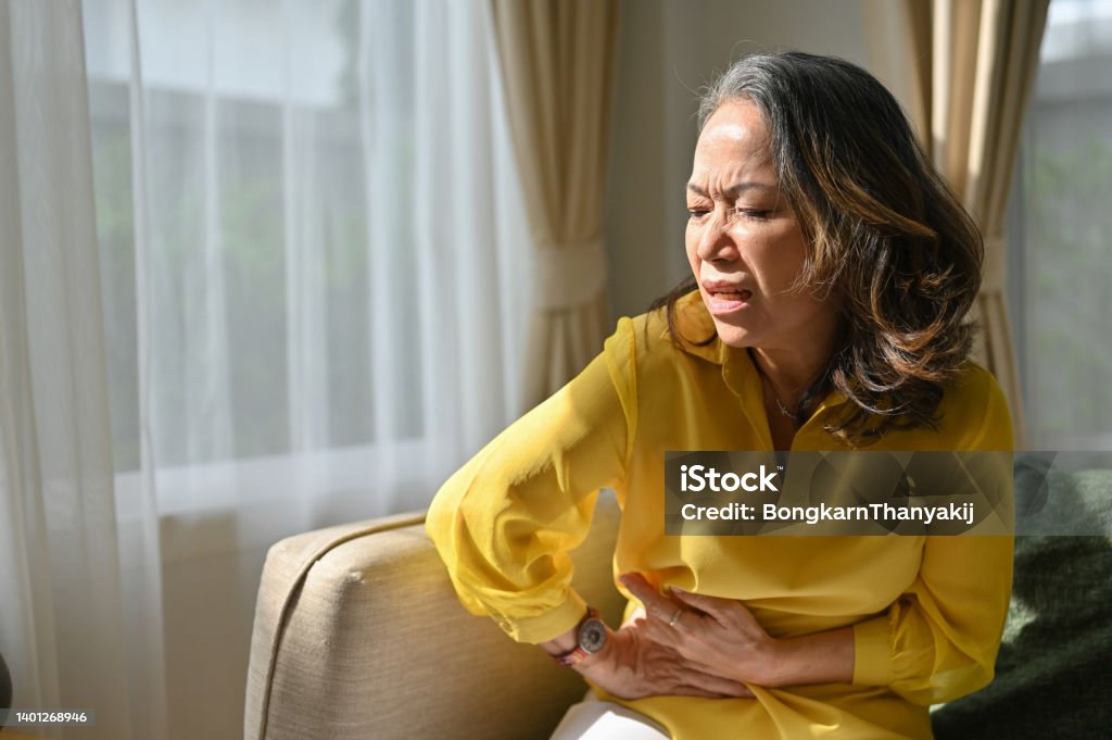 Sick asian middle aged lady hold belly suffer from abdomen ache. feeling discomfort in abdomen. Sick upset asian middle aged lady hold belly suffer from abdomen ache. Old adult woman with stomach ache, feeling discomfort in abdomen. Stomachache Stock Photo