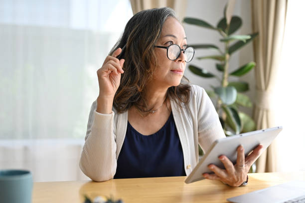 femme retraitée asiatique prospère utilisant le pavé tactile de la tablette numérique dans le salon - quinquagénaire photos et images de collection