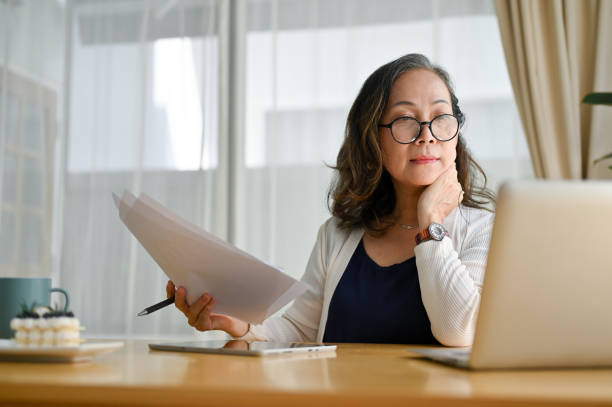 mujer de negocios asiática de mediana edad concentrada que usa una computadora portátil - looking at papers fotografías e imágenes de stock