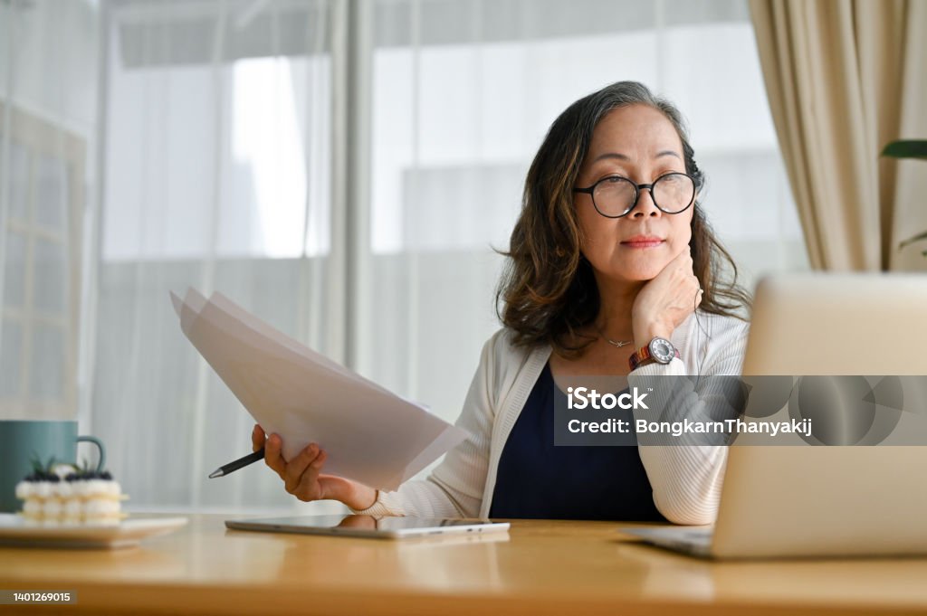 Concentrated asian middle aged female businesswoman using portable computer Concentrated asian middle aged female teacher or businesswoman in glasses sitting at desk using portable computer and examining paperwork. Age and technology Document Stock Photo