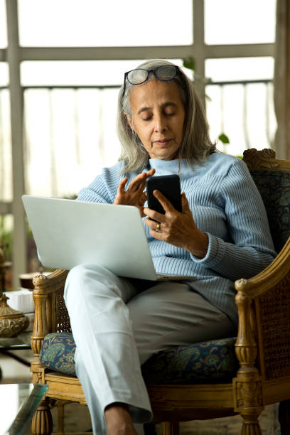 elderly woman using laptop and mobile phone at home - multi tasking asian and indian ethnicities asian ethnicity lifestyles imagens e fotografias de stock