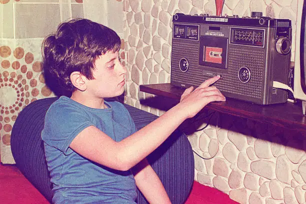 Vintage analog photo of a pre-teenage boy playing music on a stereo radio cassette player/recorder at home. Vintage image of the seventies/eighties of the 20th century.