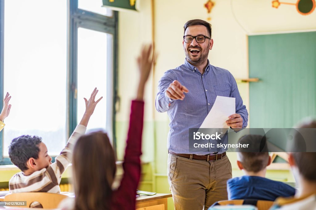 Yes you, tell me the answer to my question! Happy male teacher aiming at his students who raised their hands to answer the question on a class at elementary school. Teacher Stock Photo