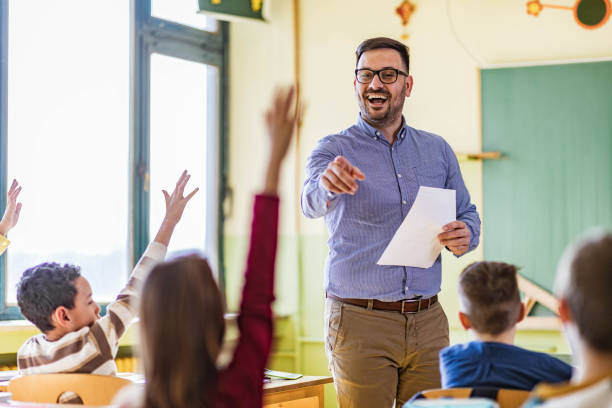 Yes you, tell me the answer to my question! Happy male teacher aiming at his students who raised their hands to answer the question on a class at elementary school. teacher stock pictures, royalty-free photos & images