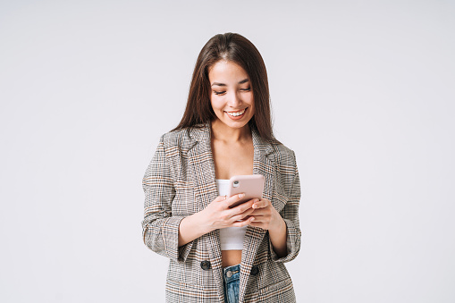 Young happy asian business woman with long hair in suit using mobile phone on grey background