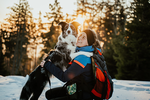 Beautiful smiling woman with backpack embracing border collie dog on snow in winter day