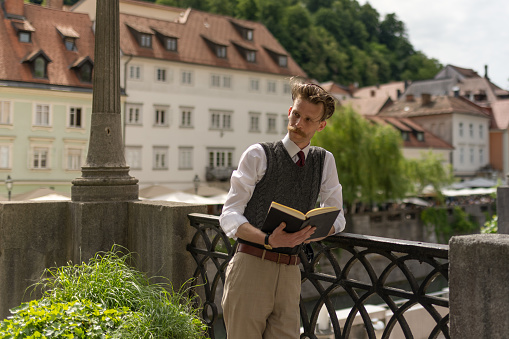 A young hipster man with a mustache on a walk around the city drinking coffee,  reading a book .