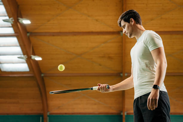 joven instructor masculino con uniforme de tenis balanceando pelota en raqueta - tennis indoors court ball fotografías e imágenes de stock