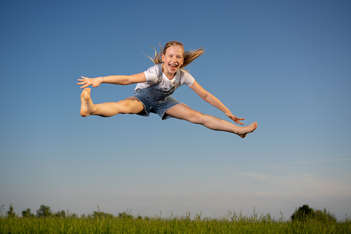Happy laughing girl jumping high on trampoline outdoors on sunny day in straddle, grass and blue sky in background