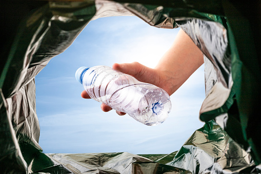 View from below of a white man hand throwing a plastic bottle on a recycle bin. Image taken from the interior of the trash bin and against the sky.