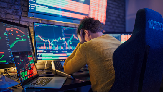 Depressed trader with head in hands sitting in front of stock market reports on computer screens at workplace