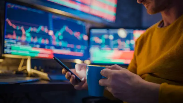 Photo of Man using mobile phone and having coffee while monitoring stock market on computer screens at desk