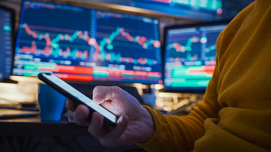 Trader text messaging on smart phone while sitting in front of stock market reports on computer screens at workplace