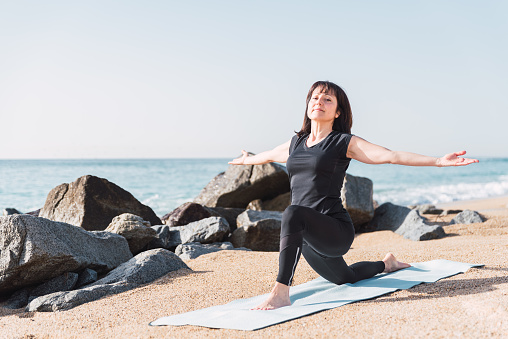 Full body of content flexible female standing in Anjaneyasana with outstretched arms on mat on sandy beach during yoga practice in summer
