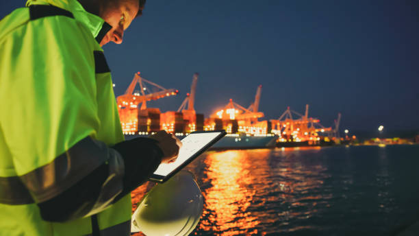 Man holding helmet and working on tablet computer in front of harbor terminal at night