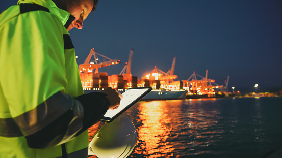 Foreperson in protective workwear using digital tablet and holding safety helmet in front of container terminal port during night