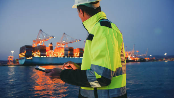 hombre con equipo de protección sosteniendo una tableta frente a la terminal del puerto por la noche - industrial ship fotos fotografías e imágenes de stock