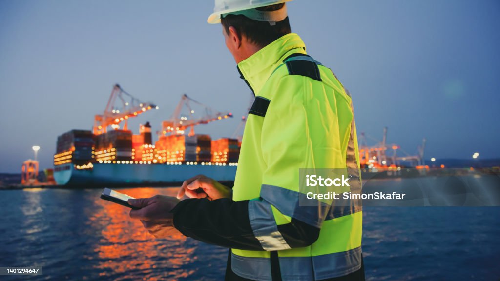 Man in protective gear holding tablet computer in front of harbor terminal at night Foreperson in protective gear using digital tablet in front of container terminal port during night Harbor Stock Photo