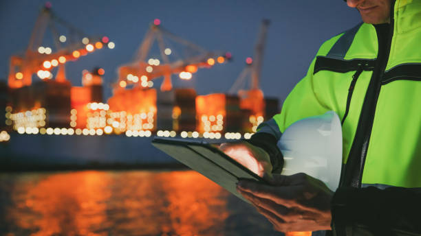 istock Man holding helmet and working on tablet computer in front of harbor terminal at night 1401294697