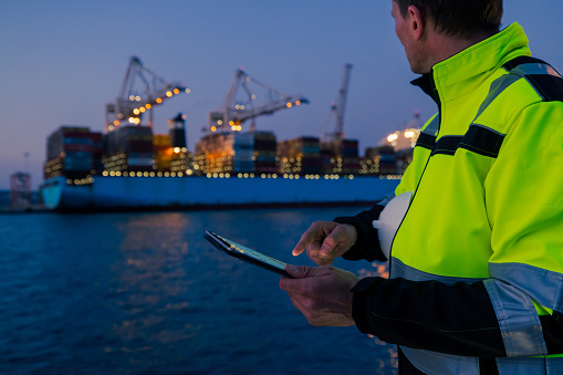 Foreperson in protective workwear using digital tablet in front of container terminal port during night