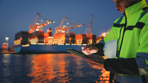 istock Man holding helmet and working on tablet computer in front of harbor terminal at night 1401295499