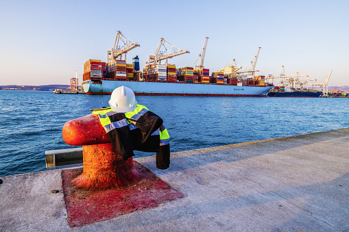 Protective gear with digital tablet placed on metal mooring bollard in front of container terminal port