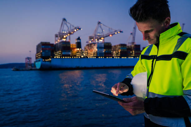 istock Man holding helmet and working on tablet computer in front of harbor terminal at night 1401295792