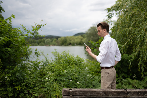 A young hipster with a mustache on a walk in a city park by the lake reads an SMS on a mobile phone and talks on a mobile phone, leaving home in a green and holy settlement.