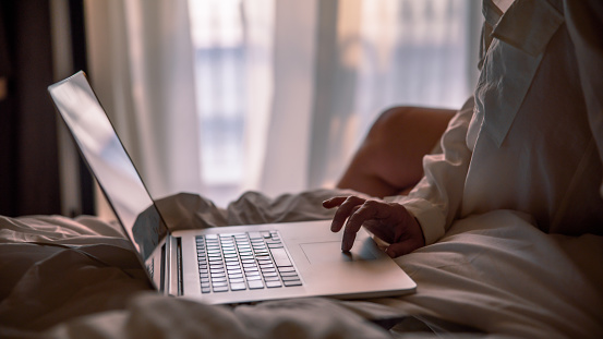 Woman in white shirt using laptop while sitting on bed in modern hotel