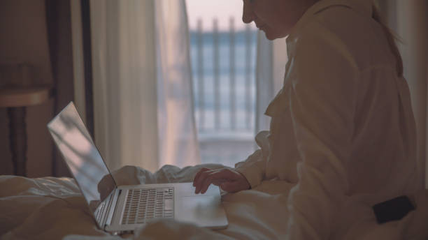 femme se relaxant sur le lit de l’hôtel et travaillant sur un ordinateur portable - curtain balcony bed sheet photos et images de collection