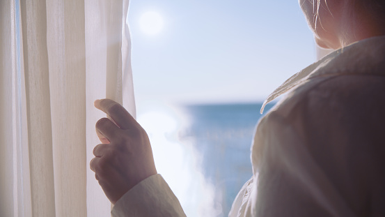 Woman in white shirt opening window curtain and enjoying sea view from balcony in modern hotel