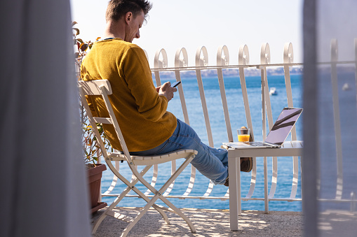 Man text messaging on smartphone while sitting beside laptop on balcony of modern hotel
