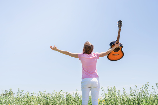 Freedom and happiness. Young woman standing in grass field raising her hand to the sky, holding guitar, view from behind
