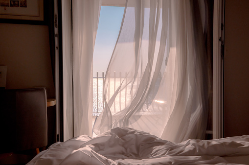 Wind moving curtains on open balcony window in modern hotel bedroom