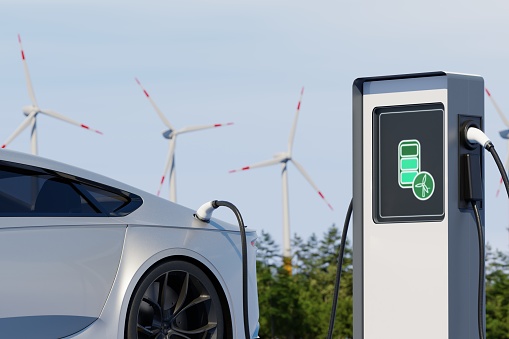 Environmentally friendly electric car charging on background of windmills. Sunny day view of EV station with port plugged in vehicle. Realistic 3d Rendering of renewable energy concept.