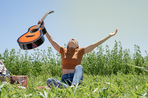 millennial woman in summer clothes and sunglasses playing guitar on a picnic. Happy woman raising her hands to the sky, holding guitar