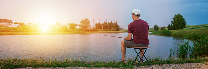 Fisher in hat seated on wooden stool standing on river bank catching fish with rod at sunset