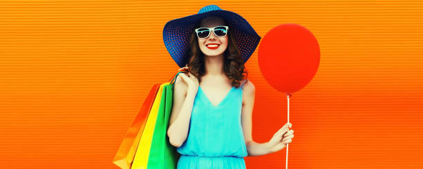 Portrait of beautiful happy smiling young woman with colorful shopping bags wearing summer straw hat on orange background, blank copy space for advertising text stock photo